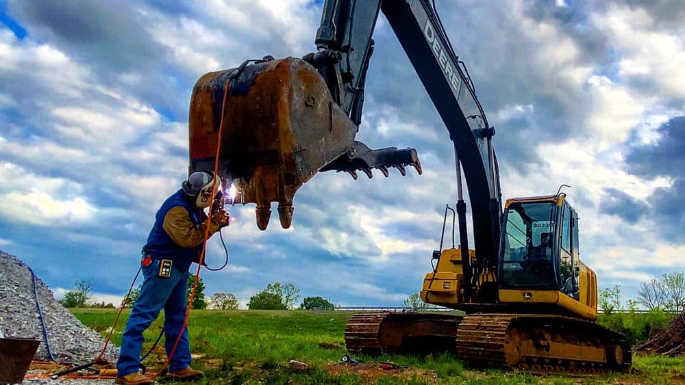 Construction worker welding on an excavator machinery under a cloudy sky.