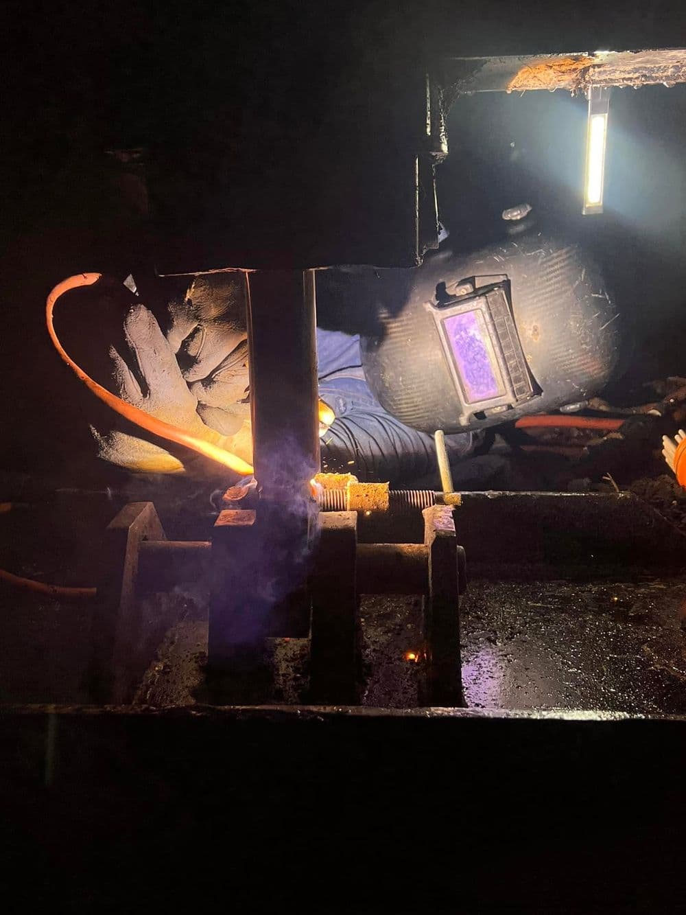 Welder working in a dark space with safety gear and equipment amid sparks and smoke.