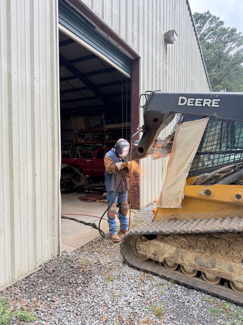 Worker welding on a John Deere skid steer loader outside a metal workshop.
