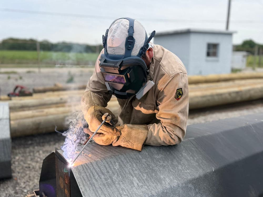 Welder in protective gear fusing metal outdoors with sparks and flames visible.